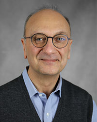 A man wearing a light blue dress shirt with a dark sweater vest and glasses.