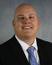 Man wearing black suit jacket with white shirt and tie against grey background.